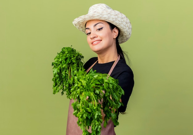 Free photo young beautiful woman gardener in apron and hat holding fresh herbs looking at front smiling cheerfully standing over light green wall