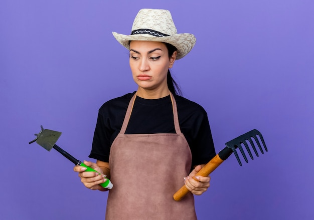 Free Photo young beautiful woman gardener in apron and hat holding gardening equipment looking confused and displeased standing over blue wall