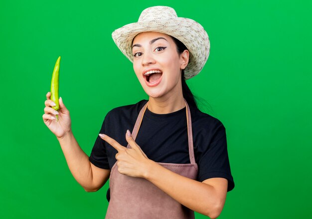 Free photo young beautiful woman gardener in apron and hat holding green chili pepper pointign with finger at it smiling standing over green wall