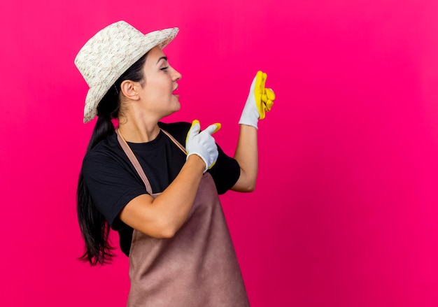 Young beautiful woman gardener in rubber gloves apron and hat pointing with index fingers back being surprised standing