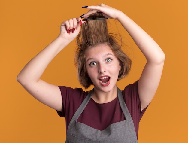 Free photo young beautiful woman hairdresser in apron combing her hair looking happy and surprised standing over orange wall