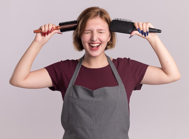 Young beautiful woman hairdresser in apron holding hair brushes near her head smiling cheerfully with closed eyes standing over white wall