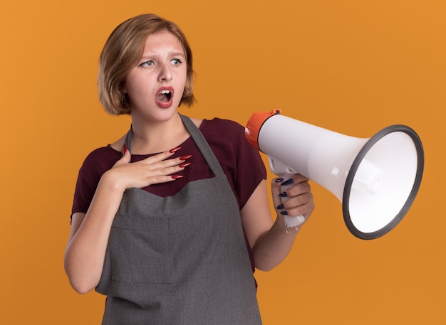 Young beautiful woman hairdresser in apron holding megaphone looking aside confused and surprised standing over orange wall