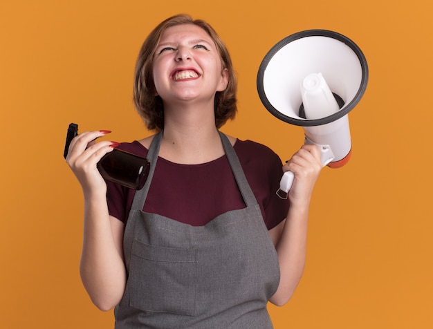 Young beautiful woman hairdresser in apron holding megaphone and spray bottle crazy happy and excited standing over orange wall