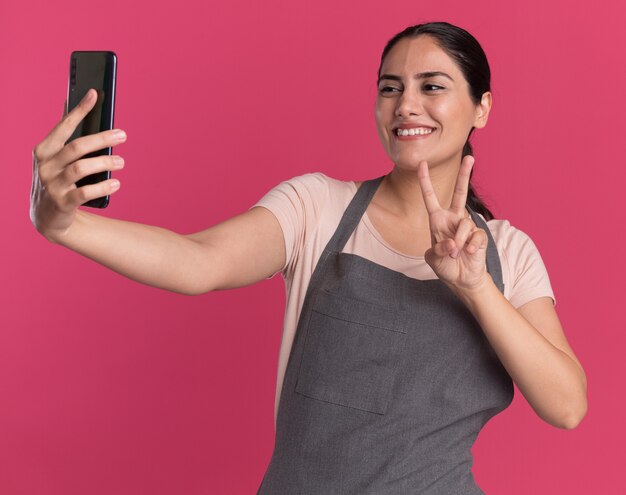 Young beautiful woman hairdresser in apron holding smartphone looking at it doing selfie smiling showing v-sign standing over pink wall