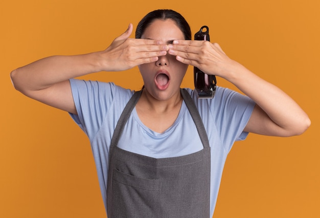 Young beautiful woman hairdresser in apron holding trimmer closing eyes with hands over orange wall