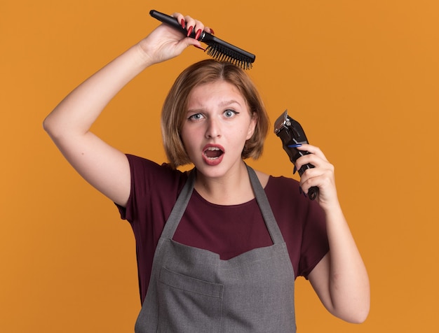 Free photo young beautiful woman hairdresser in apron holding trimmer and hair brush combing hair looking confused and surprised standing over orange wall