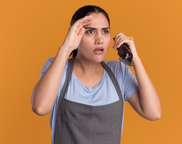 Young beautiful woman hairdresser in apron holding trimmer looking closely aside with hand over head over orange wall