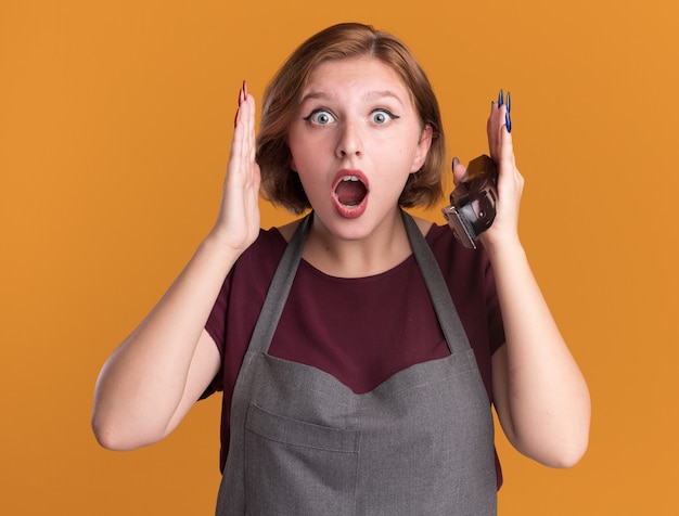 Young beautiful woman hairdresser in apron holding trimmer looking at front amazed and surprised standing over orange wall