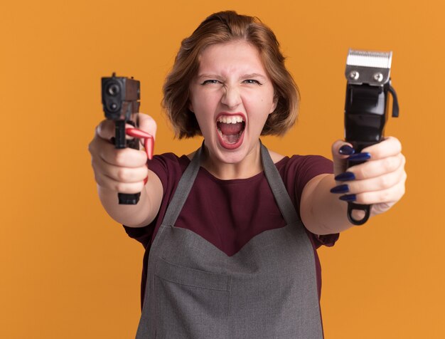 Young beautiful woman hairdresser in apron holding trimmer machine aiming with gun shouting with aggressive expression standing over orange wall