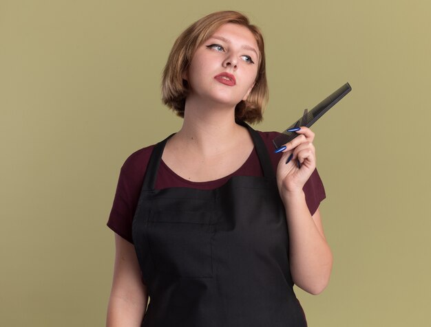 Young beautiful woman hairdresser in apron with scissors and hair comb looking aside puzzled standing over green wall