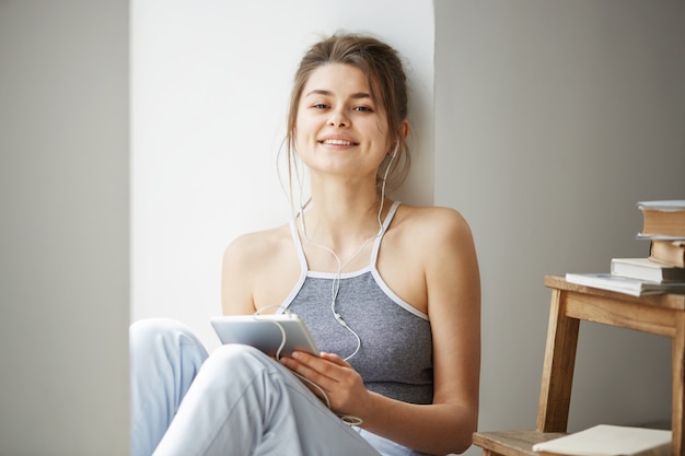 Free photo young beautiful woman in headphones smiling holding tablet listening to streaming music sitting on floor over white wall.