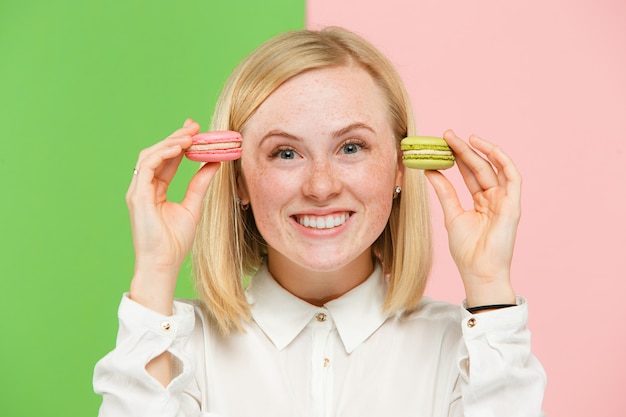 Young beautiful woman holding macaroons pastry in her hands, over trendy colored at studio.