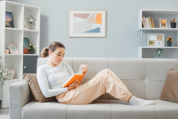 Young beautiful woman in home clothes relaxing on a couch at home interior reading a book happy and positive with smile on her face spending weekend at home