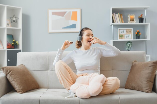 Young beautiful woman in home clothes sitting on a couch at home interior with headphones eating cookies happy and positive relaxing having fun spending weekend at home