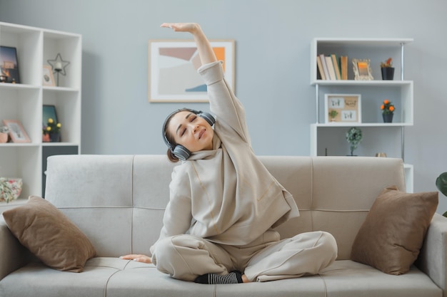 Young beautiful woman in home clothes sitting on a couch at home interior with headphones happy and positive relaxing stretching herself making exercises