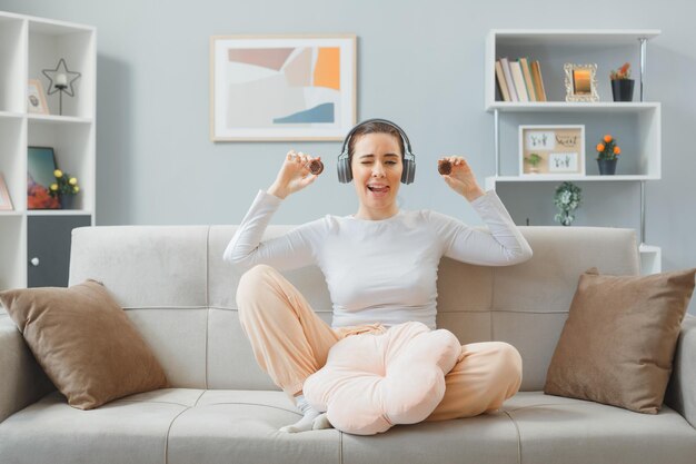 Young beautiful woman in home clothes sitting on a couch at home interior with headphones holding cookies looking at camera happy and positive sticking tongue out winking spending weekend at home