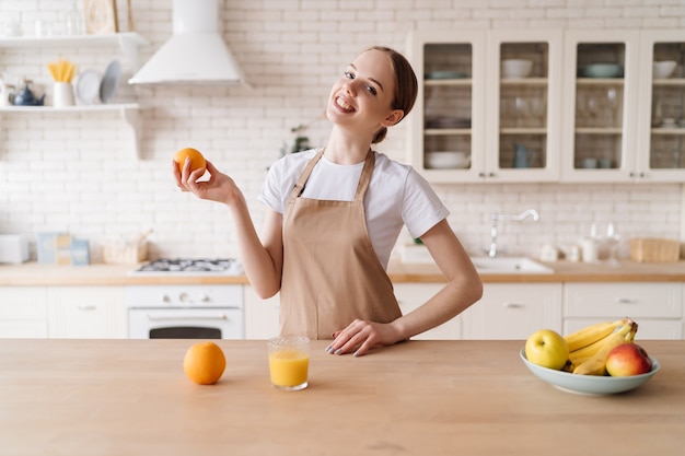 Young beautiful woman in the kitchen in an apron, fruits and orange juice