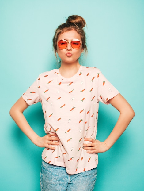 Young beautiful woman looking at camera. Trendy girl in casual summer T-shirt clothes makes duck face. Positive female shows facial emotions. Funny model isolated on blue in sunglasses