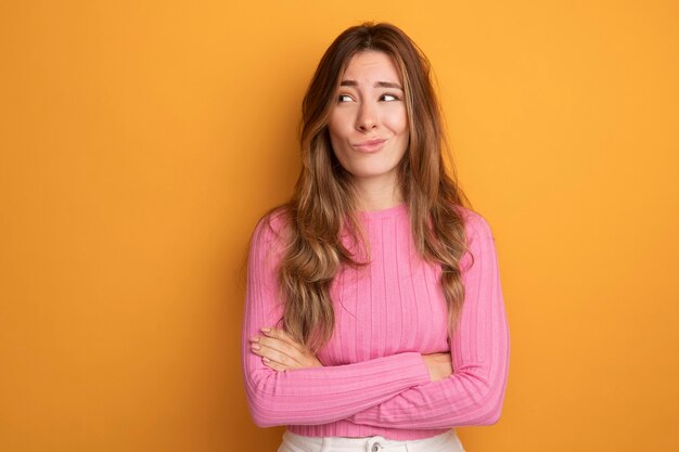 Young beautiful woman in pink top looking aside making wry mouth with disappointed expression standing over orange