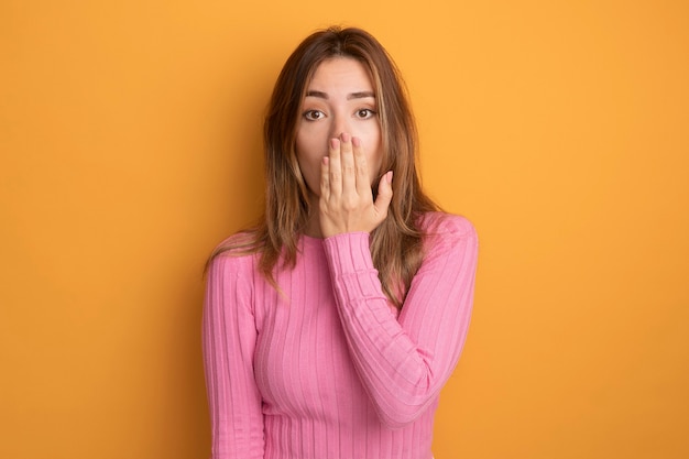 Young beautiful woman in pink top looking at camera being shocked covering mouth with hand standing over orange background