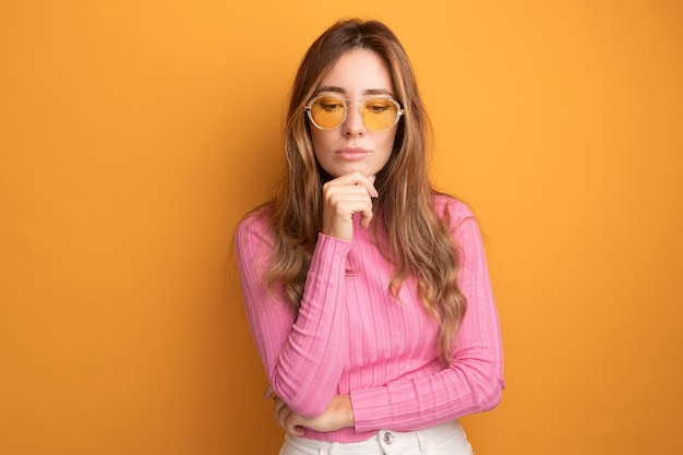 Young beautiful woman in pink top wearing glasses looking down with hand on chin with pensive expression thinking standing over orange background