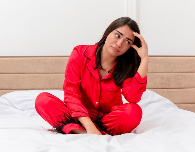 Young beautiful woman in red pajamas sitting on bed lookign aside puzzled and displeased in bedroom interior on light background