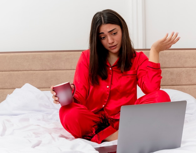 Young beautiful woman in red pajamas sitting on bed with laptop and cup of coffee looking confused and disappointed in bedroom interior on light background