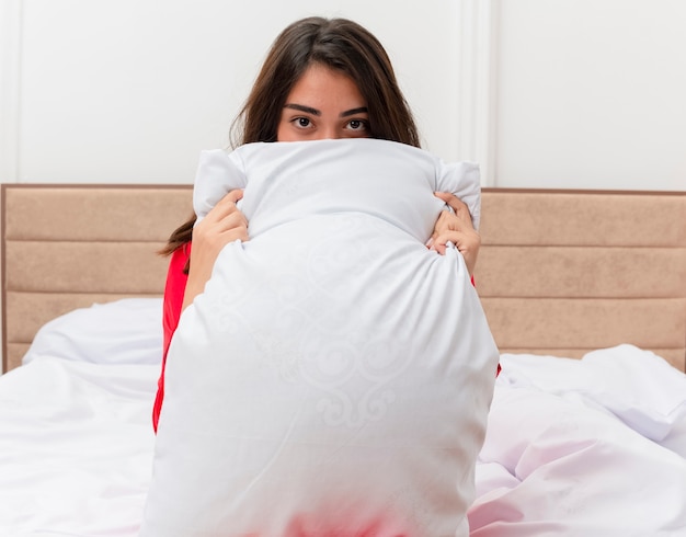 Free photo young beautiful woman in red pajamas sitting in bed with pillow hiding behind it in bedroom interior