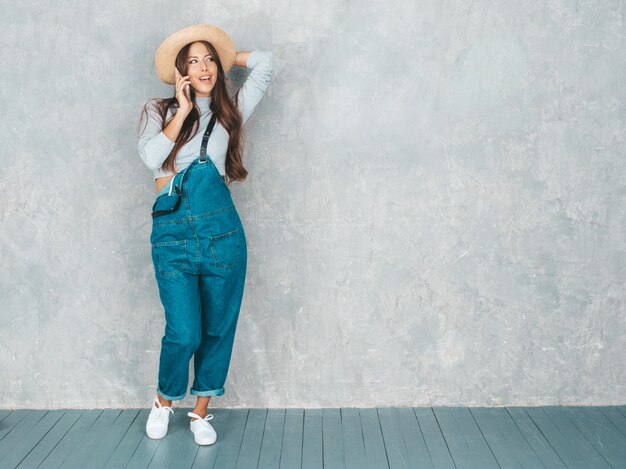 Young beautiful woman speaking on phone. Trendy girl in casual summer overalls clothes and hat. 