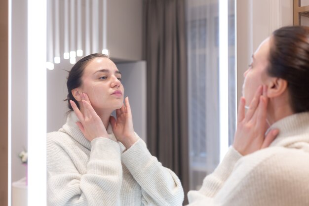 Young beautiful woman in a sweater in a beauty salon looks in the mirror, touches her face, thinks about the upcoming procedures, considers herself