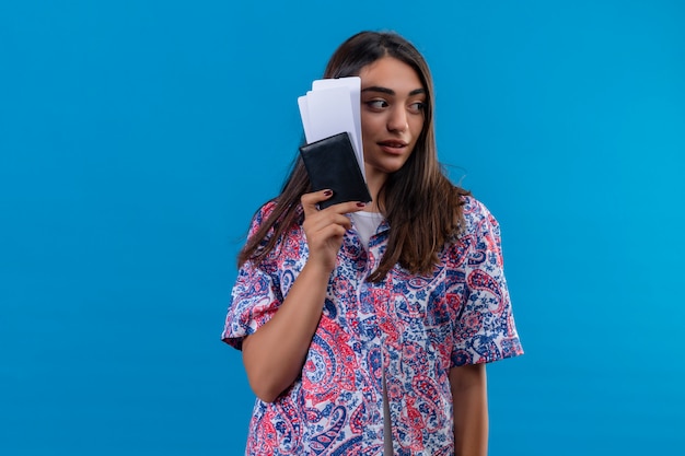 Young beautiful woman tourist holding passport with tickets looking aside with confident serious expression over isolated blue wall