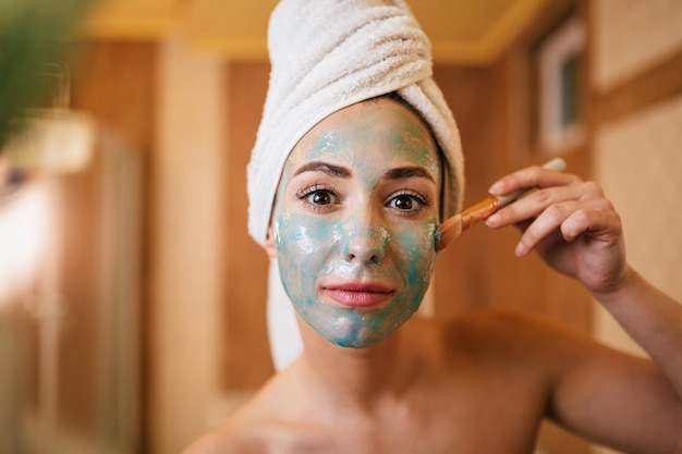 Young beautiful woman using brush while applying facial mask in the bathroom