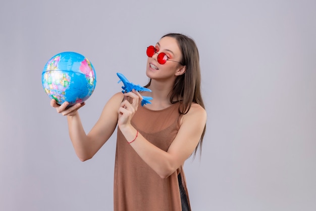 Free Photo young beautiful woman wearing red sunglasses holding globe and toy airplane playful and happy standing over white background