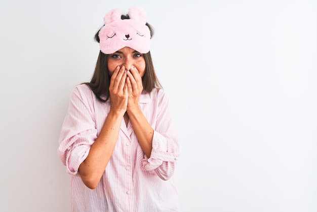 Young beautiful woman wearing sleep mask and pajama over isolated white background laughing and embarrassed giggle covering mouth with hands gossip and scandal concept