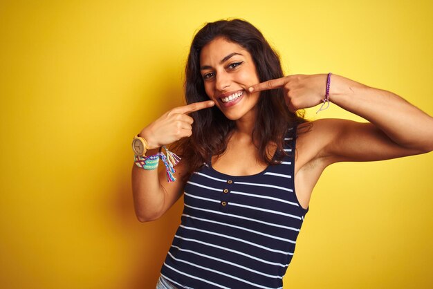 Young beautiful woman wearing striped tshirt standing over isolated yellow background smiling cheerful showing and pointing with fingers teeth and mouth Dental health concept