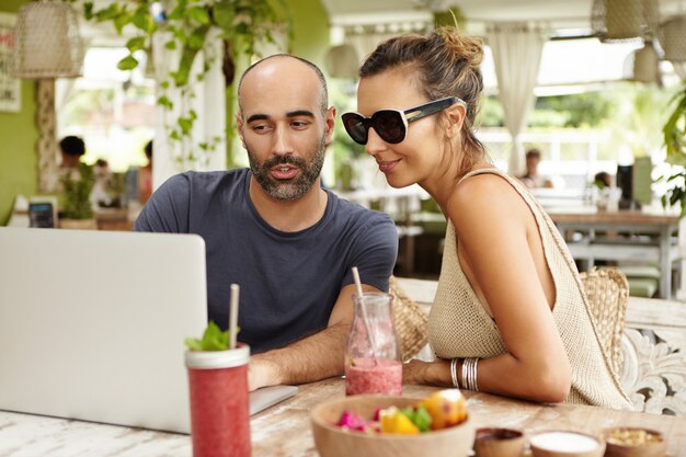 Young beautiful woman wearing sunglasses and bearded man sitting at open terrace and watching something on their generic laptop computer while browsing internet, using wireless connection during lunch