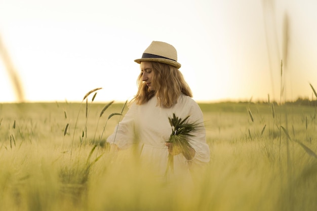 Free Photo young beautiful woman with blond long hair in a white dress in a straw hat collects flowers on a wheat field. flying hair in the sun, summer. time for dreamers, golden sunset.