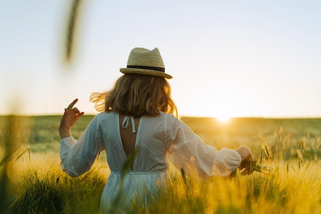 Free Photo young beautiful woman with blond long hair in a white dress in a straw hat collects flowers on a wheat field. flying hair in the sun, summer. time for dreamers, golden sunset.