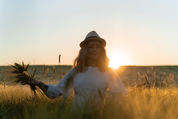 young beautiful woman with blond long hair in a white dress in a straw hat collects flowers on a wheat field. Flying hair in the sun, summer. Time for dreamers, golden sunset.