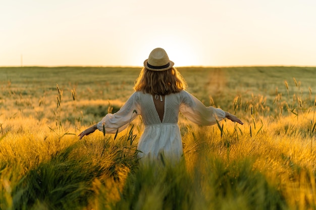 young beautiful woman with blond long hair in a white dress in a straw hat collects flowers on a wheat field. Flying hair in the sun, summer. Time for dreamers, golden sunset.