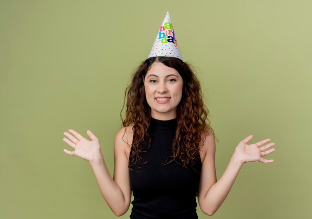 Young beautiful woman with curly hair in a holiday cap  happy and excited birthday party concept standing over light wall