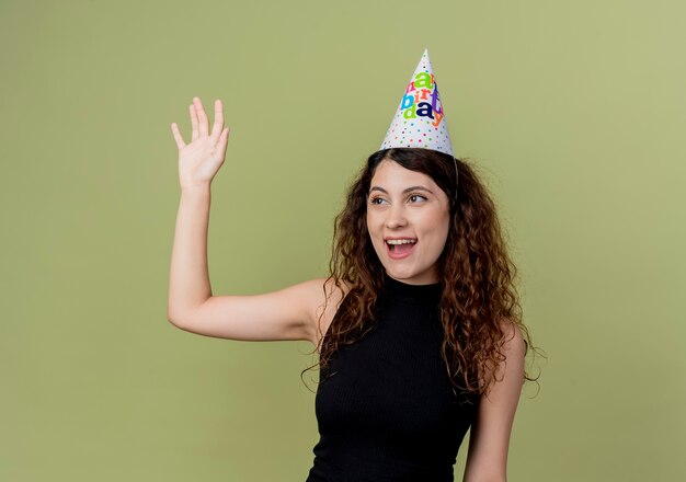 Young beautiful woman with curly hair in a holiday cap happy and positive waving with hand birthday party concept standing over light wall