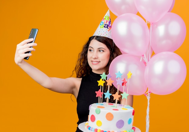 Young beautiful woman with curly hair in a holiday cap holding air balloons and birthday cake taking selfie smiling cheerfully birthday party concept standing over orange wall