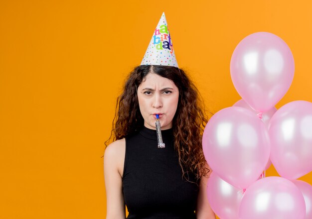 Young beautiful woman with curly hair in a holiday cap holding air balloons blowing whistle  with unhappy face birthday party concept standing over orange wall