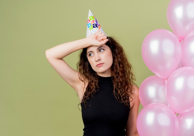 Free photo young beautiful woman with curly hair in a holiday cap holding air balloons looking tired and bored birthday party concept standing over light wall