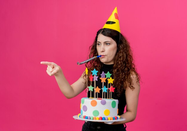 Young beautiful woman with curly hair in a holiday cap holding birthday cake blowing whistle pointing with finger to the side looking displeased birthday party concept standing over pink wall