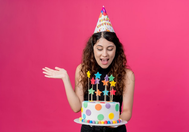 Young beautiful woman with curly hair in a holiday cap holding birthday cake happy and excited birthday party concept standing over pink wall