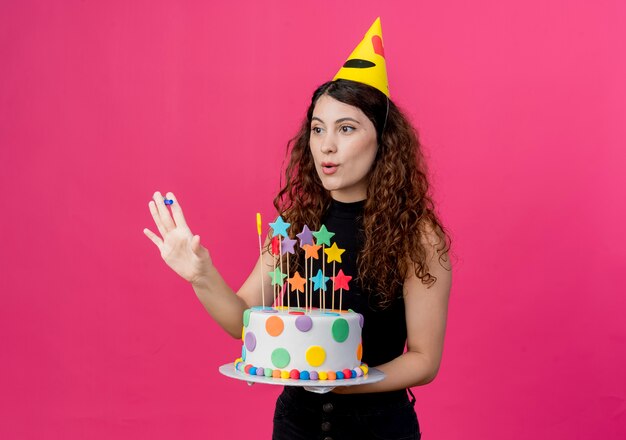 Young beautiful woman with curly hair in a holiday cap holding birthday cake happy and positive birthday party concept standing over pink wall