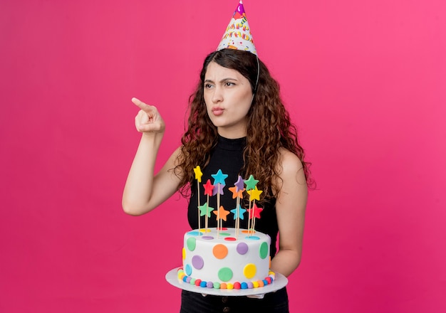 Free photo young beautiful woman with curly hair in a holiday cap holding birthday cake pointing with finger at something looking displeased birthday party concept standing over pink wall